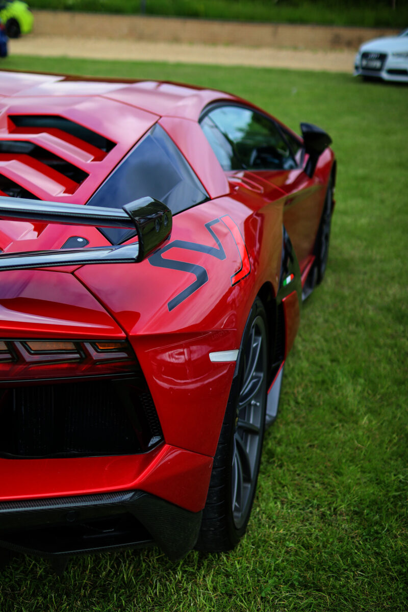 The side of a red Lamborghini Aventador SVJ on a grassy field at a car show.