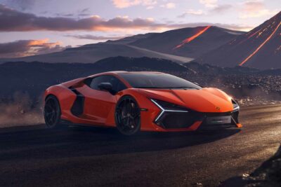 Orange Lamborghini Revuelto on a dirt road with volcanoes in the background, with the car coming to a sudden stop.