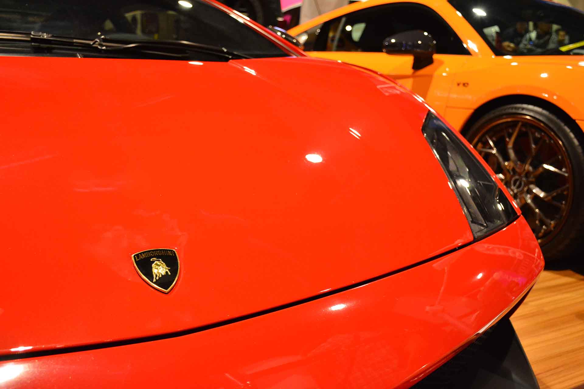The nose of an orange Lamborghini Gallardo at a car show with a Lamborghini Urus in the background.
