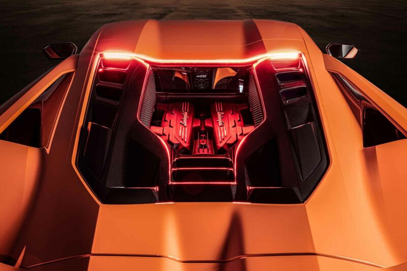 Photo of the upper brake light casting red light down onto the exposed engine of an orange Lamborghini Revuelto supercar.