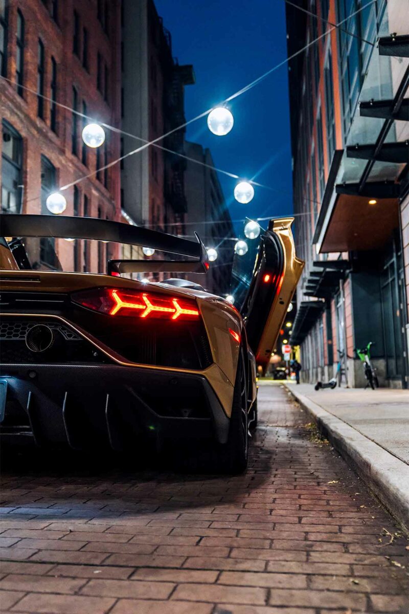 Rear end of a gold Lamborghini Aventador SVJ on a brick street at night with lights and shops in the background.