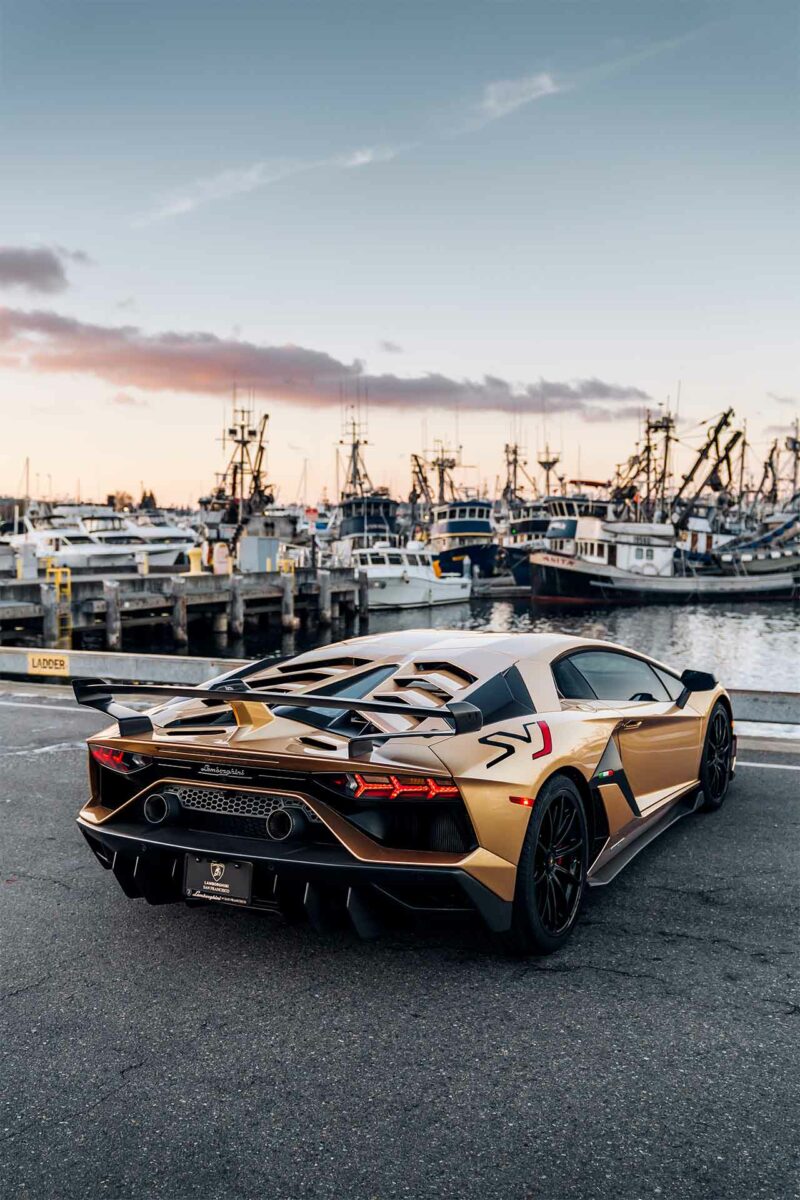 Gold Lamborghini Aventador SVJ parked in front of a dock with boats in the background at sundown.