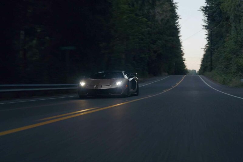 Gold Lamborghini Aventador SVJ just after sundown driving down a dark road with thick trees in the background.