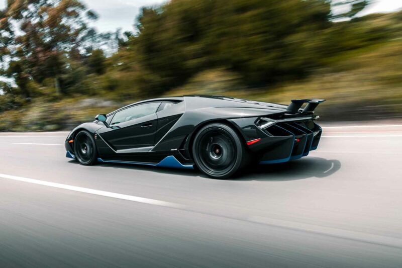 Lamborghini Centenario in black with blue accents driving on the highway with motion blurred trees in the background.